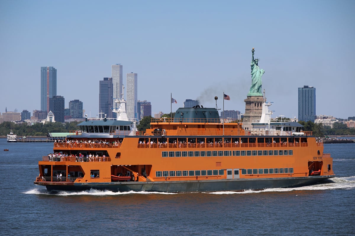 An orange ferry boat carries passengers across the blue waters of a harbor with the city skyline behind on a sunny day