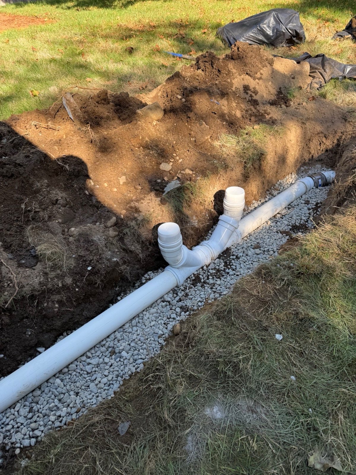Excavation trench with white PVC pipes and drainage system installed in soil with gravel bed along grass